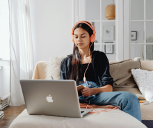 A student wearing headphones is sitting on a sofa writing in a notepad, with an open laptop in front of them.