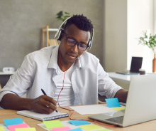 A student is sitting at a desk and writing in a notepad. They have an open laptop and scattered sticky notes in front of them,