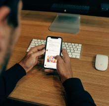 Person scrolling through social media feed on smartphone at wooden desk with keyboard and mouse nearby.