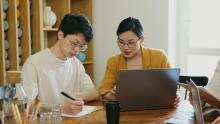 Two researchers sitting at a desk in conversation whilst typing on a laptop and writing in a notepad