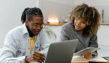 Two people are sitting next to each other at a desk. One is holding a tablet computer and resting their hand on a book. The other is typing on an open laptop.