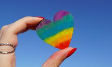 Close-up of a hand holding a multicoloured heart ornament, representing LGBT+ pride colours.