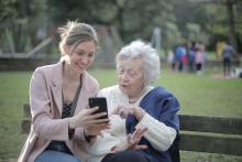Young woman showing something on her smartphone to an elderly woman sitting together on a park bench during daytime.