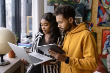 Two students in a home environment are looking at an open laptop, with one student holding a tablet computer
