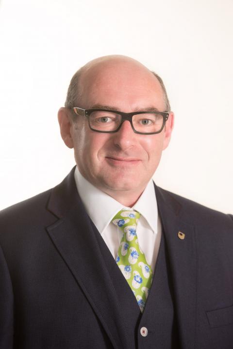 A picture of John D'Arcy who is wearing glasses and a suit and green tie