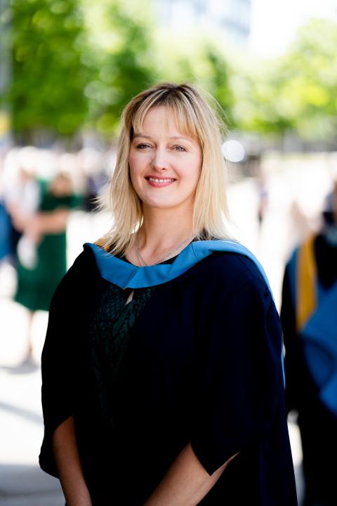 blonde haired woman wears her graduation robes and smiles into the camera