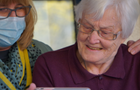 An woman with a mask on putting her hands on the shoulder of an elderly woman as she smiles