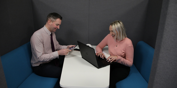 A man and woman sat at a desk working and laughing on a phone and laptop