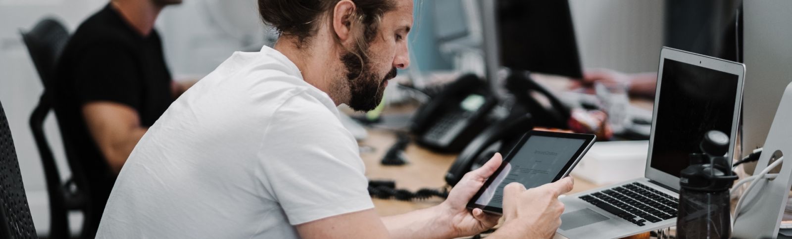 a male with a beard, wearing a white t-shirt sits at a desk looking at an ipad