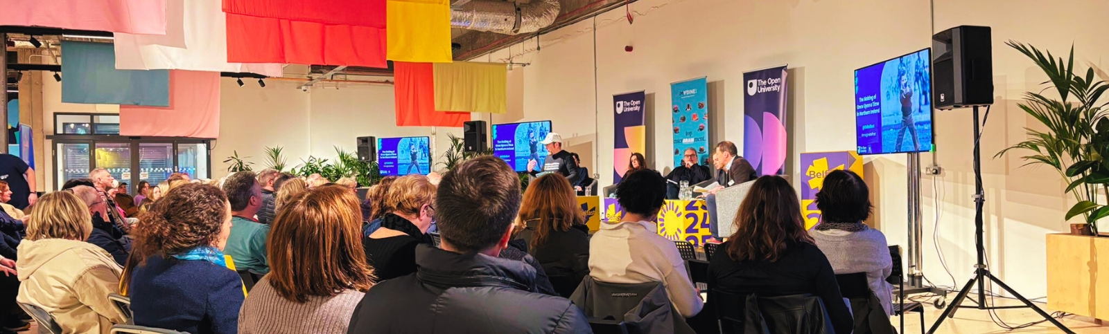 a crowd of people watching an event which has a man standing talking at the front and a panel of people seated behind him