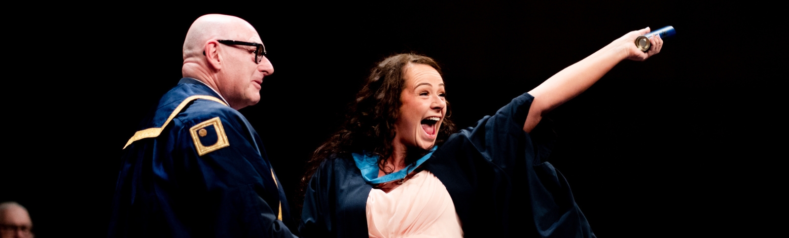 a female graduate with black hair, smiling in her graduation gown with a bald man with thick black glasses and wearing a graduation robe