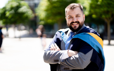 Jason outside in OU degree ceremony robing. He's got his arms folded and is smiling into the camera