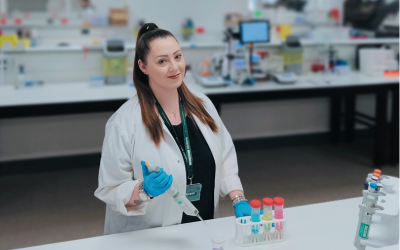 Jenny is in a lab wearing a lab coat and gloves handling a syringe