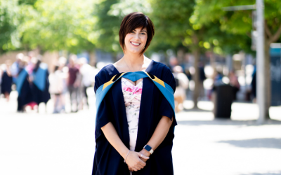 Katie is stood outside in the sun smiling into the camera while wearing OU degree ceremony robes.