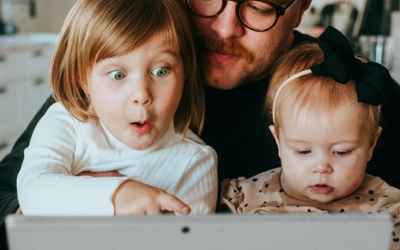 a man sits with two children on his knee as they look at a laptop in front of them