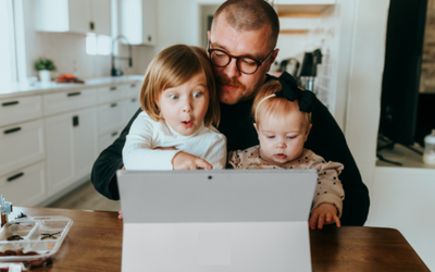 A man with two young girls on his knee pointing with excited faces towards a laptop on the counter.