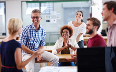 A diverse group of people sitting at and on a desk smiling and laughing with a woman slightly out of focus