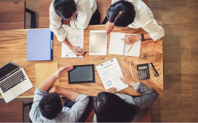 A top down view of a wooden table with four people working over documents, a tablet and a laptop.