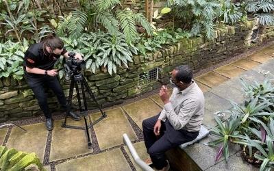 A man sits talking into a camera, they are wearing facemasks