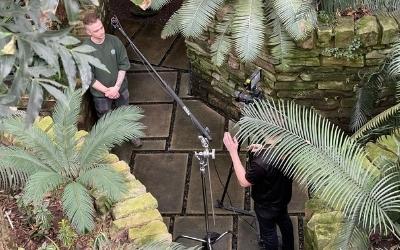 A man is being filmed talking into a camera surrounded by plants