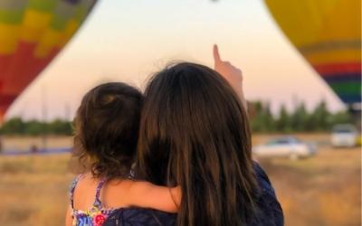 a woman holds a child in her arms. they are facing away from the us and pointing up a hot air balloon