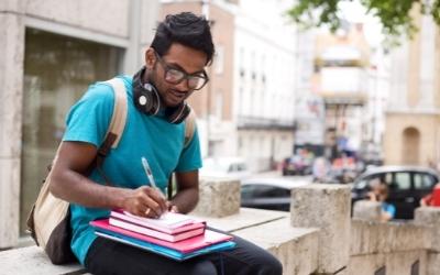 a man in a turquoise t-shirt sits on a wall with books on his knee and he writes in a notebook 