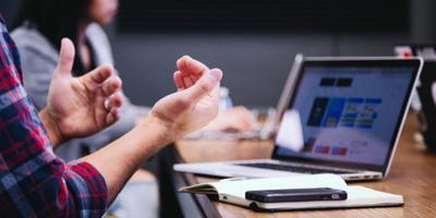 a mans hands gesturing as he talks seated a a desk with a laptop in front of him and a notebook and mobile phone