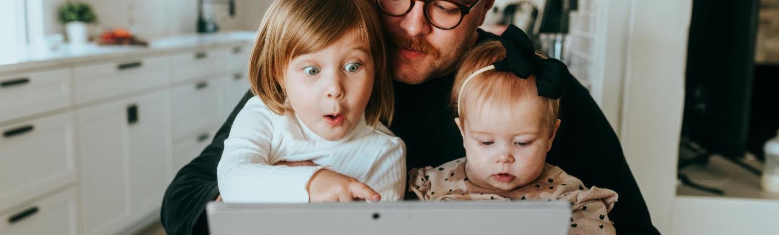 a father sits at a table with his two children on his knee looking at a laptop