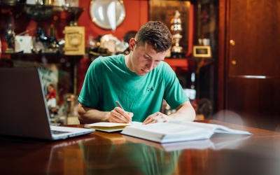 A young man wearing a green tshirt studying at a desk writing in a notepad with a laptop in front of him