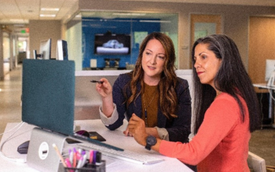 two women look at a laptop together at a desk, one holds a pen as she points at the screen