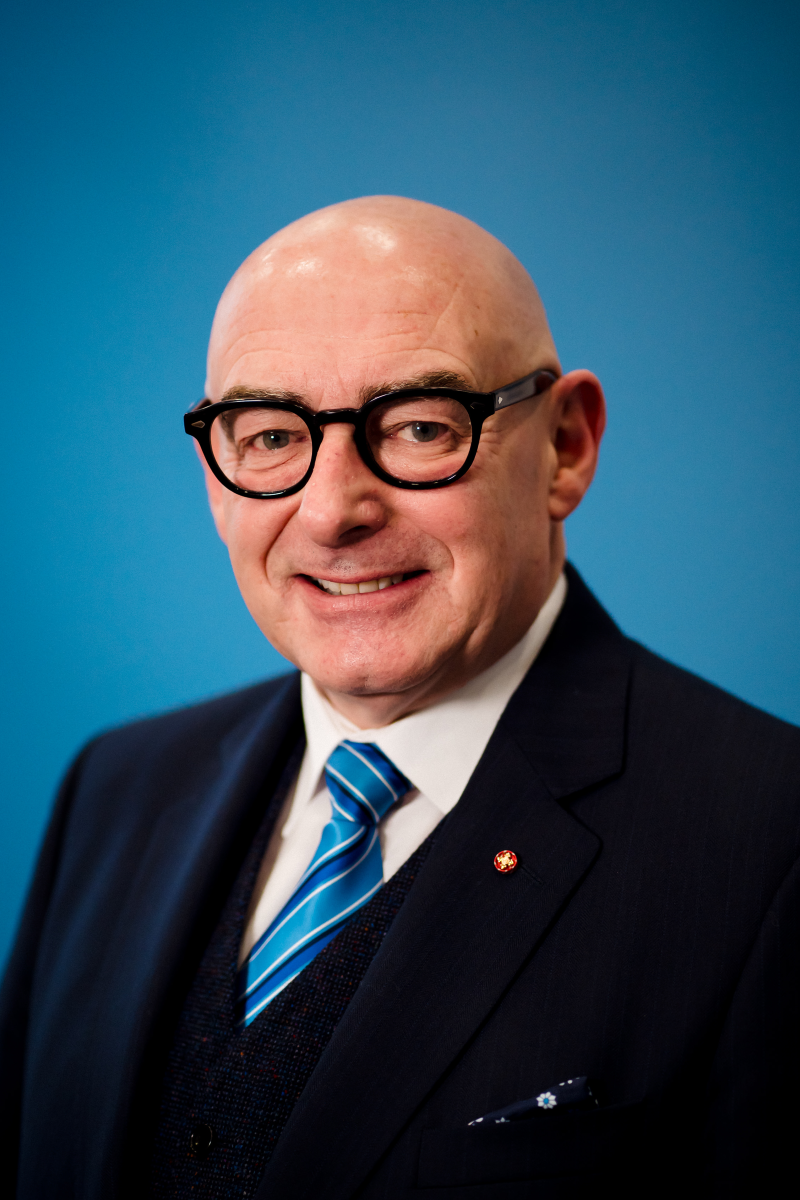 A picture of John D'Arcy, he is bald and has thick black glasses and is smiling wearing and suit and tie