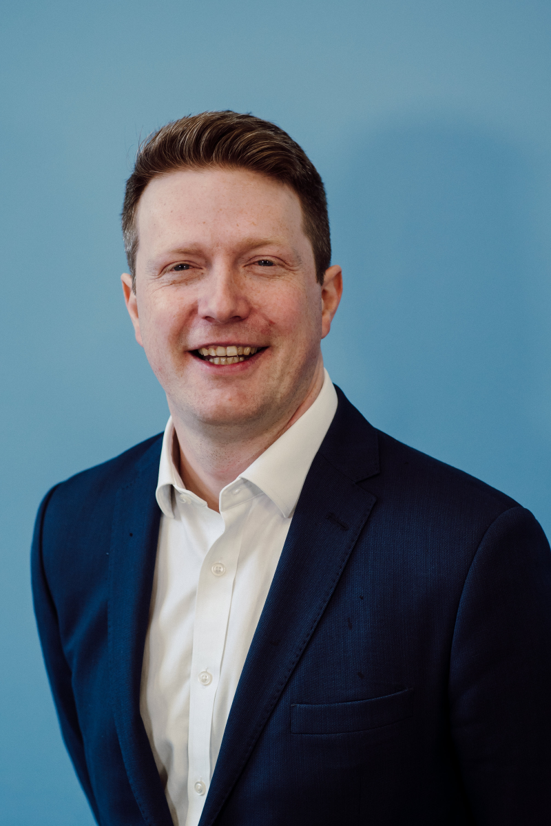 A picture of Michael bower who has fair short hair and is smiling and wearing a suit