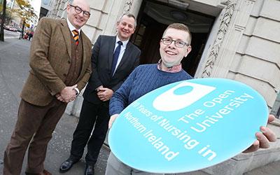 three men pose for a photo, one is holding a board with the OU logo afn words saying 10 years of Nursing in Northern Ireland