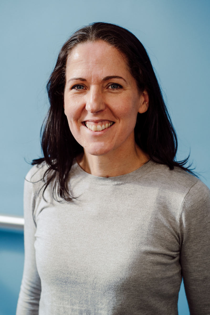 A picture of Tara Craig who has dark hair and is smiling wearing a grey sweater