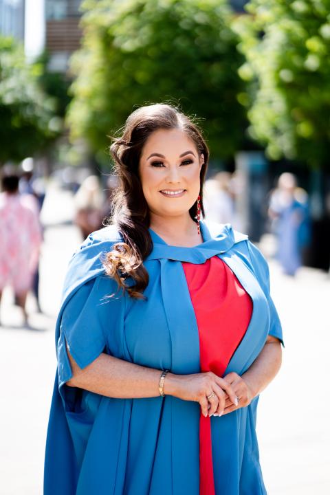 a woman stands in her graduation robes, she has long dark hair and smiles into the camera