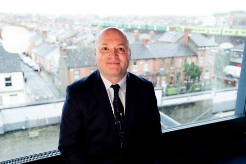 Brian, dressed in a suit, sat smiling to camera in front of a window onto a housing in Dublin.