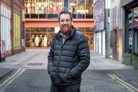 Callum is smiling to camera with his hands in his puffer jacket pockets. He's stood on a high street in belfast.