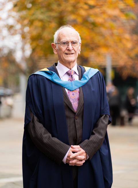 An elderly man wearing graduation robes and a suit stands outdoors, smiling proudly. The background shows autumn trees with orange leaves, creating a warm and celebratory atmosphere.