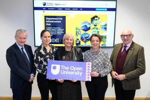 Five people stand smiling in front of a screen displaying The Open University’s OpenLearn “Department for Infrastructure Hub” webpage. The group holds a purple sign with The Open University logo. The image represents the collaboration between The Open University and the Department for Infrastructure to launch a new online learning hub.