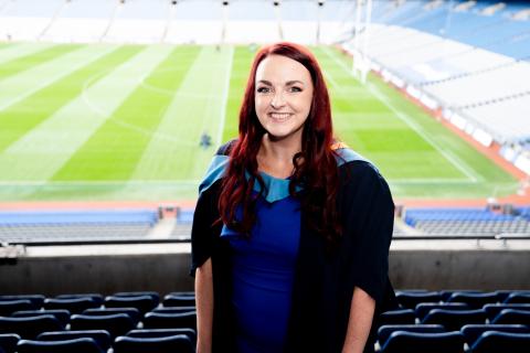 A smiling woman with long red hair wearing a blue dress and black graduation gown stands in a stadium, with green pitch and empty seats in the background.