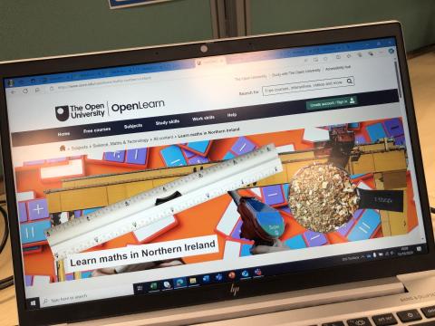 A computer monitor displaying The Open University OpenLearn webpage. The website features a colorful graphic with images of mathematical tools, including a ruler, measuring tape, and measuring spoon. The webpage title reads "Learn maths in Northern Ireland," and a search bar is visible at the top along with navigation tabs for subjects, free courses, and study skills.