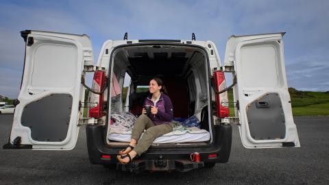 Hailee, a woman in her early 20s, sits at the back of a van with its doors open, enjoying a moment of relaxation. She is holding a mug and wearing casual outdoor clothing, including a purple jacket and khaki pants. The van is parked in a scenic area with green hills in the background. She is smiling away from the camera while holding a mug.