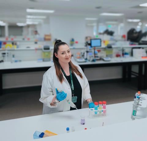 a woman stands in a lab with a lab coat on and is working with some equipment on the desk in front of her