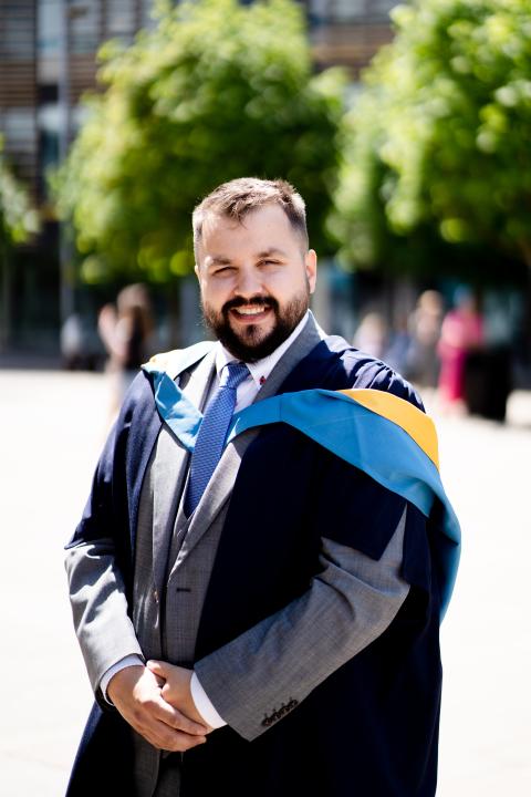 A picture of Jason in his graduation robes. He has dark hair and a beard and is smiling