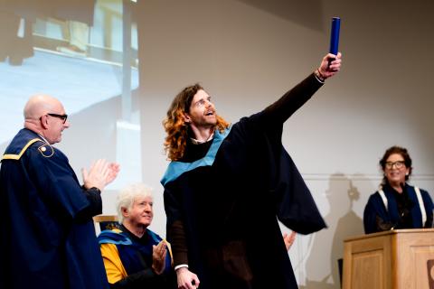 Jonathan crossing the stage at an OU degree ceremony celebrating with his scroll held out towards the crowd off screen.