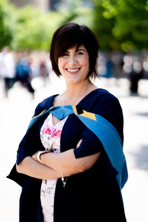 Katie is dressed in her graduation robes.  She has short dark hair and smiles into the camera