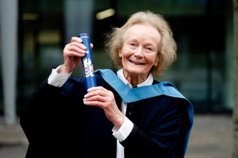 Am photo of Maryette in Open University degree ceremony robes hold a scroll and smiling to camera.