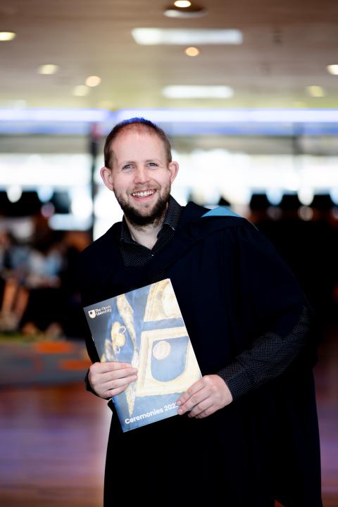 Michael stands in his graduation robes holding a graduation programme