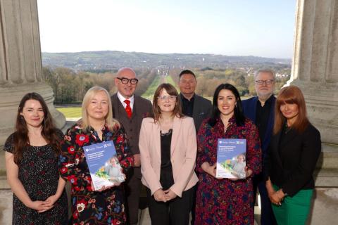 Minister for the Economy Dr Caoimhe Archibald stands with members of the Alliance for Lifelong Learning on the terrace at Parliament Buildings, Stormont. The group is posed smiling in front of scenic parkland, with two members holding copies of a report titled "Measuring the Impact of Adult Education in Northern Ireland."