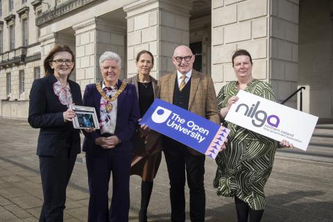 A group of five people stand outside a grand stone building, smiling at the camera. Two individuals hold signs displaying "The Open University" and "NILGA - Northern Ireland Local Government Association," while another holds a tablet showing an online learning platform. One person wears a ceremonial mayoral chain, signifying a leadership role. The image represents the launch of a new learning hub initiative for local councillors in Northern Ireland.